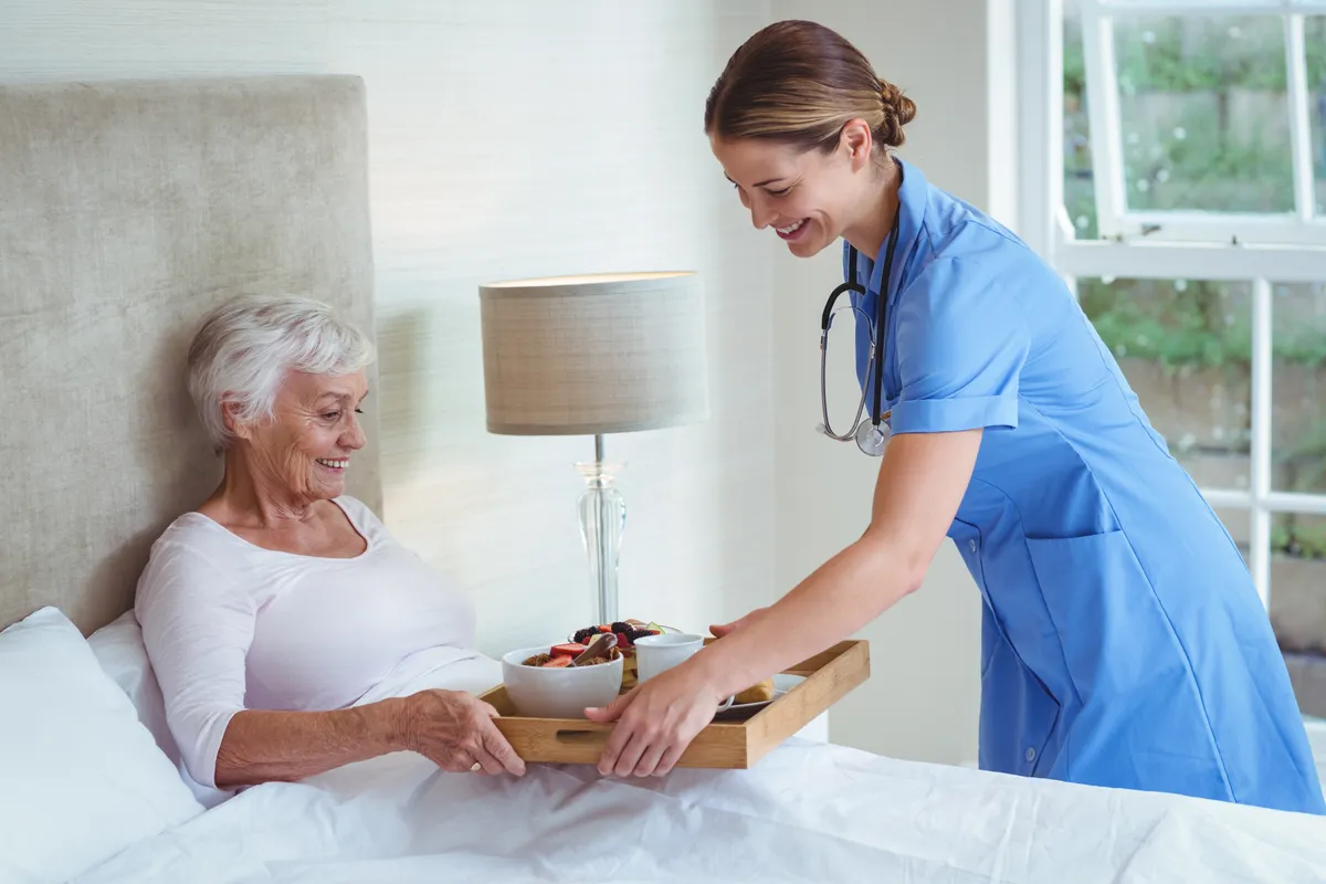 A caregiver bringing a warm breakfast to a smiling client at home